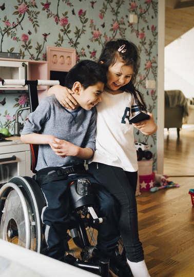 Young girl hugging a boy in a wheelchair, both looking at a mobile phone