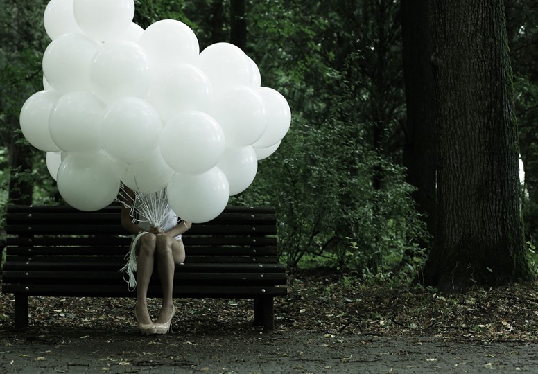 Girl sat on bench with head obscured by bunch of white balloons