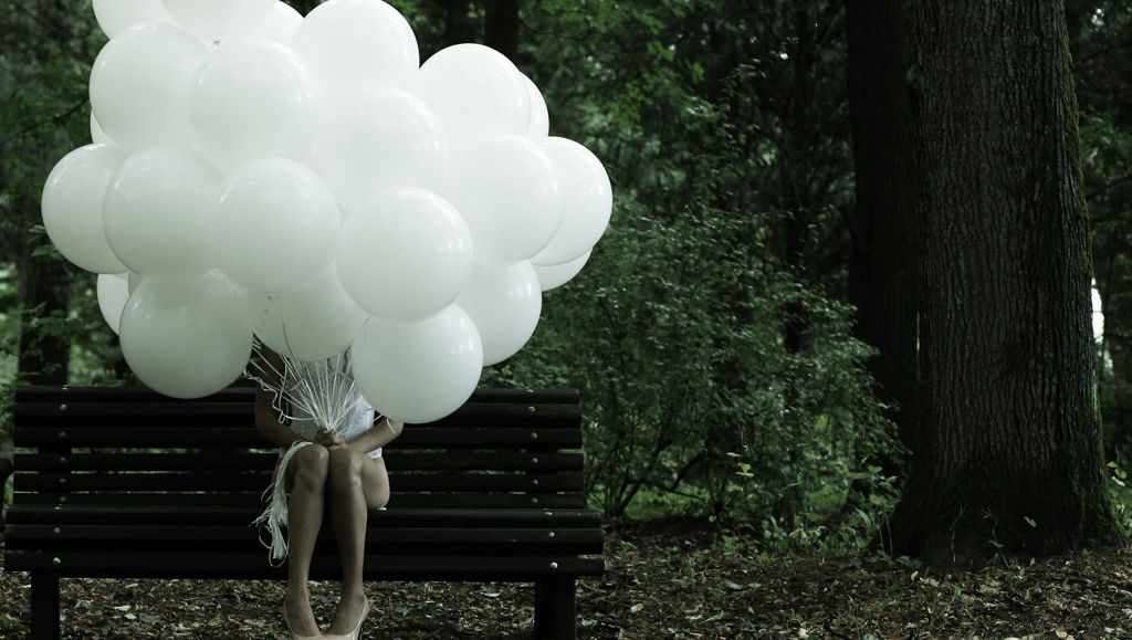 Girl sat on bench with head obscured by bunch of white balloons