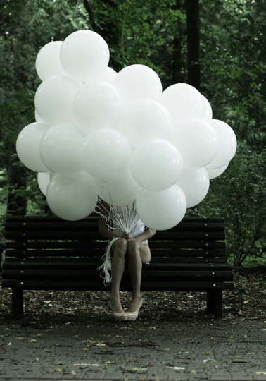 Girl sat on bench with head obscured by bunch of white balloons