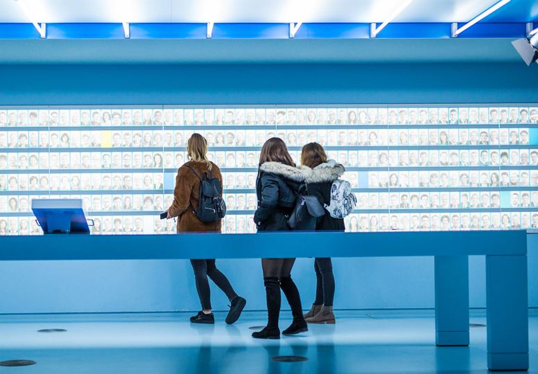 Three young girls looking at hundreds of images of faces on a wall